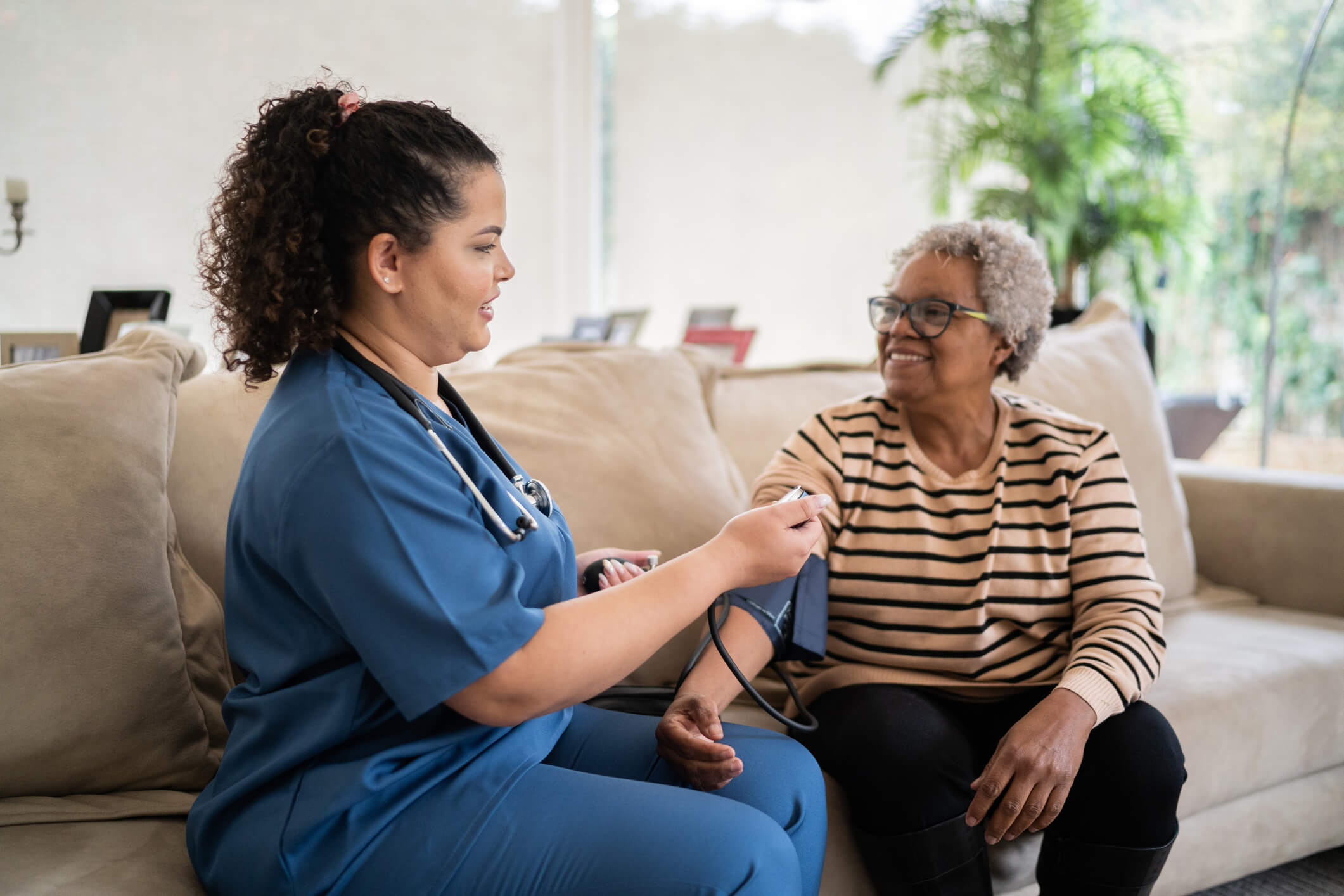 A nurse takes a blood pressure reading of a senior woman. They are in a house sitting on a couch.